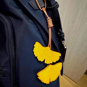 May include: A dark navy blue backpack featuring a decorative charm made of two bright yellow ginkgo leaf-shaped cutouts. The leaves are attached to the bag by a thin, light brown leather cord that is knotted at the top and loops through a black strap.