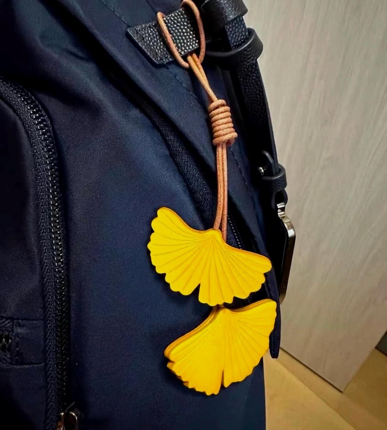 May include: A dark navy blue backpack featuring a decorative charm made of two bright yellow ginkgo leaf-shaped cutouts. The leaves are attached to the bag by a thin, light brown leather cord that is knotted at the top and loops through a black strap.
