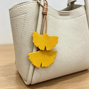 May include: A cream-colored handbag with a textured surface and two top handles. Attached to the handle is a decorative charm featuring two bright yellow ginkgo leaf shapes suspended from a thin, light brown cord tied in a simple knot.
