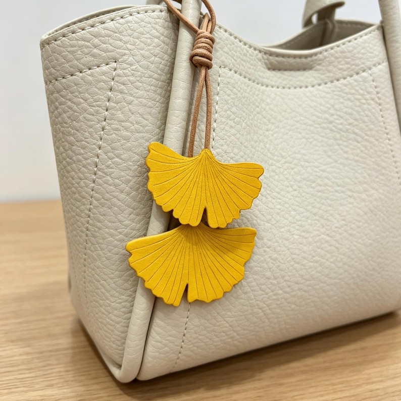 May include: A cream-colored handbag with a textured surface and two top handles. Attached to the handle is a decorative charm featuring two bright yellow ginkgo leaf shapes suspended from a thin, light brown cord tied in a simple knot.