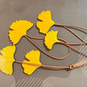 May include: Four bright yellow ginkgo leaf-shaped ornaments attached to thin, light brown leather cords. The cords are gathered together at one end with a wrapped leather detail, creating a decorative arrangement of fan-shaped foliage elements.