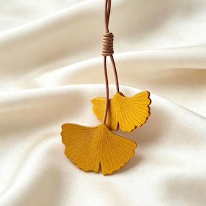 Può includere: Un ciondolo con due elementi gialli a forma di foglia di ginkgo sospesi a un sottile cordino marrone. Le foglie presentano una finitura opaca con sottili linee incise a raggiera. Il cordino è annodato in alto, fissando le foglie in posizione.