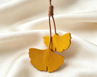 May include: A pendant featuring two yellow ginkgo leaf-shaped charms suspended from a thin brown cord. The leaves have a matte finish with fine, radiating etched lines. The cord is knotted at the top, securing the two leaves in a dangling position.