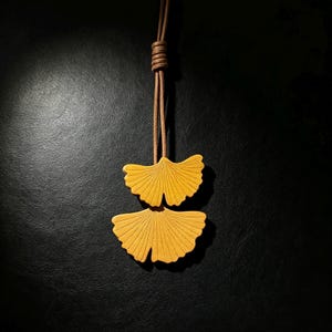 May include: A necklace featuring two yellow ginkgo leaf pendants suspended from a brown leather cord. The leaves have a textured surface with radiating vein lines. The cord is knotted at the top, creating a simple and clean design for this accessory.