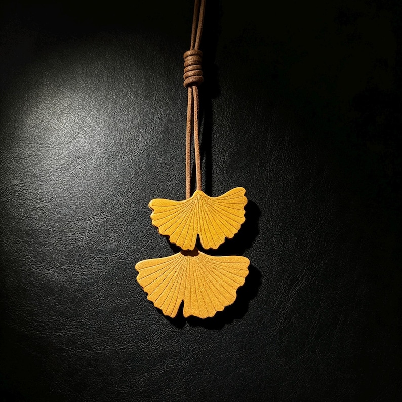 May include: A necklace featuring two yellow ginkgo leaf pendants suspended from a brown leather cord. The leaves have a textured surface with radiating vein lines. The cord is knotted at the top, creating a simple and clean design for this accessory.