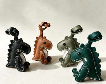 May include: Four dinosaur-shaped keychains made of leather in black, tan, olive green, and dark green. Each keychain features white contrast stitching, a small black eye, a white belly detail, and a looped leather strap at the top for attachment.