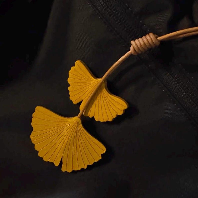 May include: A necklace featuring two bright yellow ginkgo leaf pendants attached to a tan leather cord. The leaves have a distinct fan shape with fine, radiating vein lines etched into the surface. A small, coiled leather knot secures the cord.
