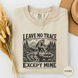 May include: An ivory t-shirt featuring a black graphic print of a Bigfoot walking through a forest with a mountain in the background. The text reads LEAVE NO TRACE EXCEPT MINE and MOUNT HOOD OREGON. A small star charm hangs from the neckline.