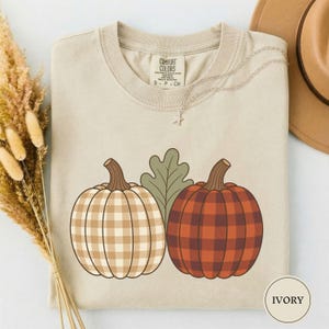May include: A beige crewneck t-shirt featuring a graphic of two pumpkins with a central green leaf. One pumpkin has a tan gingham pattern, and the other has a red and black buffalo plaid pattern. The shirt has a Comfort Colors brand label.