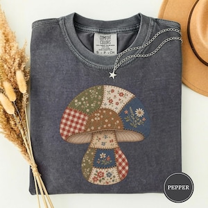 May include: A charcoal gray short-sleeve t-shirt featuring a large mushroom graphic composed of various patchwork patterns including floral prints and red gingham. The shirt has a crew neckline and a Comfort Colors label at the inner collar.