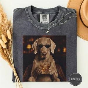 Op de afbeelding: Een antracietgrijs t-shirt met ronde hals en een grafische print van een hond met een zonnebril die een glas met amberkleurige vloeistof vasthoudt. Het shirt heeft een Comfort Colors-label in de nek en het woord PEPPER in een cirkel onderaan.