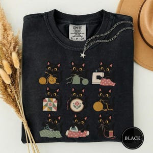 Könnte beinhalten: Ein schwarzes T-Shirt mit Rundhalsausschnitt und einem Grafikdruck von neun schwarzen Katzen, die verschiedenen Handarbeitstätigkeiten wie Stricken, Nähen und Sticken nachgehen. Das Shirt hat ein Comfort Colors Etikett und eine silberne Sternkette.