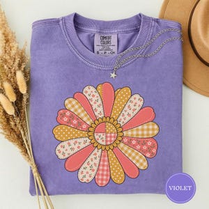 May include: A violet short-sleeve t-shirt featuring a large, central graphic of a flower with petals in alternating patterns of pink floral, yellow polka dot, and orange gingham. The shirt has a crew neckline and a Comfort Colors brand label.