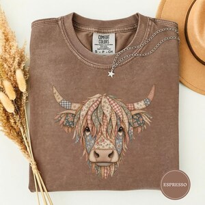 Pode incluir: Uma t-shirt castanha de gola redonda com um gráfico de uma vaca Highland composto por vários padrões florais e xadrez. A peça tem uma etiqueta Comfort Colors no pescoço e um colar com um pendente de estrela prateado na gola.