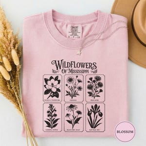 May include: A light pink crewneck t-shirt featuring a black graphic print titled Wildflowers of Mississippi. The design includes six rectangular panels displaying botanical illustrations of a magnolia, coreopsis, wild aster, cardinal flower, black-eyed susan, and blue flag iris.
