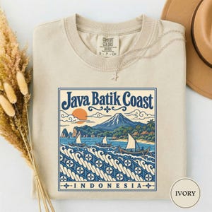 May include: An ivory t-shirt featuring a graphic print with the text Java Batik Coast Indonesia. The design depicts a mountain, palm trees, and sailboats on water with a stylized wave pattern. A thin silver necklace rests on the neckline.