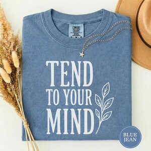 Puede incluir: Una camiseta azul de manga corta con el texto blanco TEND TO YOUR MIND y un gráfico de hoja. La camiseta presenta una etiqueta Comfort Colors en el cuello interior y un collar plateado con un pequeño dije de estrella sobre la tela.