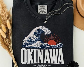 May include: A black crewneck t-shirt featuring a graphic of a stylized ocean wave and a red sun with rays. Below the graphic, the text reads OKINAWA JAPAN in white capital letters. The shirt has a Comfort Colors label at the inner neckline.