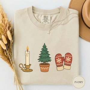 Puede incluir: Una camiseta de cuello redondo color marfil con un estampado gráfico de una vela encendida en un soporte dorado, un pequeño árbol de hoja perenne en maceta y un par de manoplas rojas con patrones de copos de nieve blancos. Etiqueta Comfort Colors.