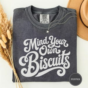 Op de afbeelding: Een antracietgrijs t-shirt met korte mouwen en een vintage wassing. Het shirt is voorzien van de witte tekst Mind Your Own Biscuits in een vetgedrukt cursief lettertype. Een zilverkleurige ketting met een sterretje ligt op de halslijn.