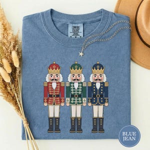 May include: A blue short-sleeved t-shirt featuring a graphic of three nutcracker figures in red, green, and blue uniforms with gold crowns. The shirt has a Comfort Colors label at the neck and a silver star-shaped pendant necklace resting on it.