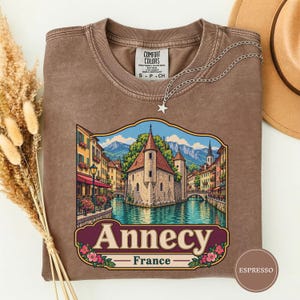 Op de afbeelding: Een bruin t-shirt met ronde hals en een grafische print van een stenen gebouw aan een kanaal in Annecy, Frankrijk. Het ontwerp bevat kleurrijke bloemen, een bergachtergrond en de tekst "Annecy France" in een decoratief kader op de borst.