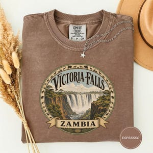May include: A brown short-sleeve t-shirt with a circular graphic featuring Victoria Falls in Zambia. The design includes a border of small animal silhouettes and the text Victoria Falls and Zambia. The shirt has a Comfort Colors brand label.