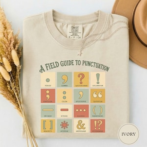 May include: An ivory t-shirt featuring a graphic grid titled A FIELD GUIDE TO PUNCTUATION. The grid displays sixteen colorful squares, each containing a specific punctuation mark and its corresponding name, such as period, comma, and ampersand.