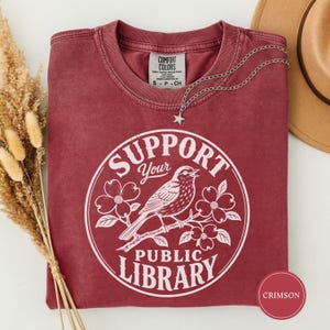 Puede incluir: Una camiseta de cuello redondo color carmesí con un gráfico circular blanco de un pájaro posado en una rama florida con el texto Support Your Public Library. La prenda tiene una etiqueta Comfort Colors y un collar con dije de estrella.