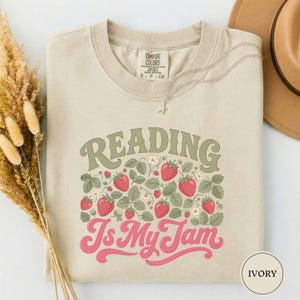 Puede incluir: Una camiseta de cuello redondo color marfil con el texto "READING Is My Jam" en verde salvia y rosa. El diseño incluye un gráfico de fresas rojas y hojas verdes. La camiseta tiene una etiqueta Comfort Colors en el cuello interior.