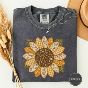 May include: A charcoal gray crew neck t-shirt featuring a large sunflower graphic composed of various patterned fabric-style petals in yellow, orange, and white, including gingham and floral prints. The shirt has a Comfort Colors brand label.