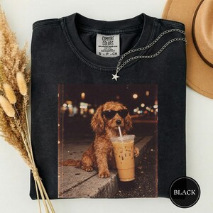Puede incluir: Una camiseta negra de cuello redondo con un estampado gráfico de un perro marrón con gafas de sol negras que sostiene un vaso de plástico con pajita. La camiseta tiene una etiqueta Comfort Colors y un collar de cadena plateada con estrella.
