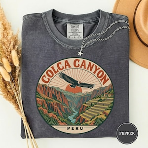 Peut inclure: Un t-shirt à col rond gris anthracite avec un motif circulaire du canyon de Colca au Pérou. Le design montre un condor en vol, une rivière, des terrasses et un soleil. Le texte indique Colca Canyon et Peru. Un collier étoile argenté est inclus.