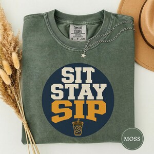 May include: A moss green crewneck t-shirt featuring a dark blue circular graphic with the text SIT STAY SIP in white and gold block letters. Below the text is a simple line drawing of a drink cup with a straw. The shirt has a Comfort Colors label.