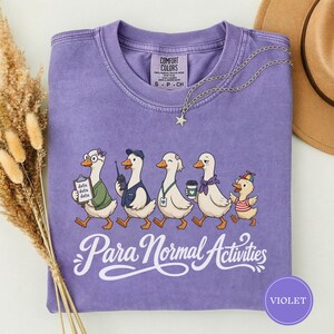 Puede incluir: Una camiseta violeta de manga corta con un gráfico de cinco patos y el texto "Para Normal Activities". El primer pato lleva gafas y sostiene un portapapeles con "data data data", el segundo una gorra, el tercero un cordón, el cuarto una bufanda y el quinto un sombrero pequeño.