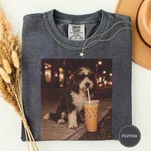 Op de afbeelding: Een antracietgrijs t-shirt met ronde hals en een grafische print van een pluizige hond met een zonnebril, zittend naast een ijskoffie met een rietje. Het t-shirt heeft een Comfort Colors-label bij de hals en een ster-vormige ketting.