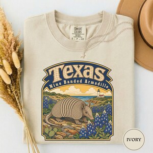 May include: An ivory t-shirt featuring a graphic print of a nine-banded armadillo in a field of bluebonnets near a lighthouse and the ocean. The text reads Texas and Nine Banded Armadillo. The shirt has a crew neckline and a Comfort Colors label.