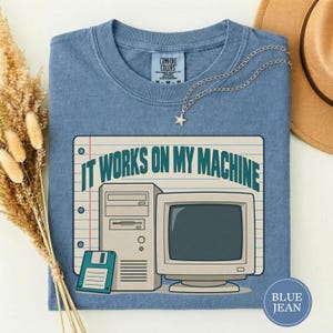 May include: A blue crew neck t-shirt featuring a graphic of a vintage computer tower, a monitor, and a floppy disk on a lined paper background. The text IT WORKS ON MY MACHINE is printed in dark teal above the computer equipment illustration.