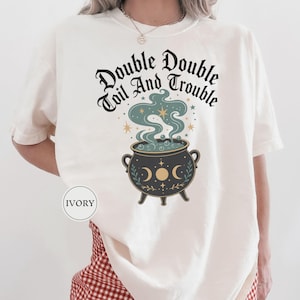 May include: An ivory t-shirt featuring a black graphic of a cauldron with steam rising from it. The text "Double Double Toil And Trouble" is printed in a black gothic font above the cauldron, which is decorated with moon phases and stars.