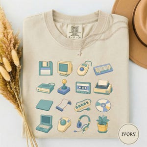 May include: A beige crew neck t-shirt featuring a grid of retro computer and office technology illustrations, including a floppy disk, monitor, mouse, keyboard, joystick, cassette tape, printer, power strip, CD, headphones, and a potted plant.