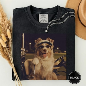 May include: A black t-shirt featuring a graphic print of a dog wearing a visor and sunglasses while sitting in a golf cart. The dog has its paw on a golf club. The shirt has a Comfort Colors label at the neck and a silver star necklace.