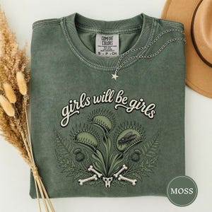 Może przedstawiać: Mchowo-zielony t-shirt z okrągłym dekoltem i napisem "girls will be girls" nad grafiką przedstawiającą muchołówki, paprocie, osty i kości. Na kołnierzyku koszulki spoczywa srebrny łańcuszek z małą zawieszką w kształcie gwiazdki.