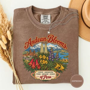 Peut inclure: Un t-shirt marron à manches courtes avec une impression graphique de montagnes andines, de fleurs et de bâtiments. Le texte indique Andean Blooms, Cusco Arequipa Puno Ancash Junin of Peru. Le t-shirt a un col rond et une étiquette Comfort Colors.