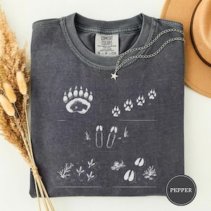 Op de afbeelding: Een antracietgrijs t-shirt met witte grafische prints van diverse diersporen, waaronder een berenpoot, hoefafdrukken van een hert en vogelpootafdrukken. Het t-shirt heeft een ronde hals en een Comfort Colors-label in de kraag. Een zilveren sterrenketting ligt op de stof.