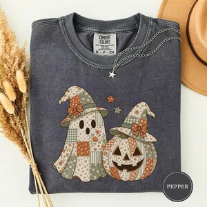 Könnte beinhalten: Ein anthrazitfarbenes T-Shirt mit Rundhalsausschnitt und einem Aufdruck eines Geistes sowie eines Kürbisses, die beide spitze Hüte mit floralem Patchwork- und Karomuster tragen. Das T-Shirt hat ein Comfort Colors-Etikett am inneren Halsausschnitt.