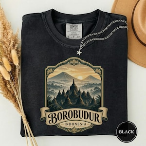 May include: A black crewneck t-shirt featuring a graphic print of the Borobudur temple in Indonesia. The design is framed in an ornate border with the text "BOROBUDUR INDONESIA" on a banner. The shirt has a Comfort Colors brand label inside.
