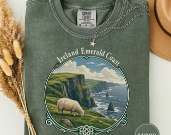May include: A moss green crewneck t-shirt featuring a graphic of a sheep grazing on a cliffside overlooking the ocean. The text reads Ireland Emerald Coast. A silver star pendant necklace rests on the neckline of the shirt.