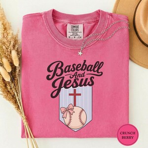 Puede incluir: Una camiseta rosa de manga corta con un estampado gráfico que incluye el texto Baseball And Jesus sobre una cruz roja y una pelota de béisbol con un lazo rosa. La camiseta tiene cuello redondo y una etiqueta de la marca Comfort Colors.
