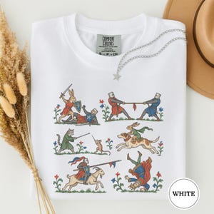 以下が含まれることがあります： 中世風のイラストがプリントされた白いTシャツ。騎士と戦うウサギ、ネズミとフェンシングをする猫、犬に乗るウサギなど、動物や騎士が様々なシーンで描かれたグラフィックが特徴で、Comfort Colorsのラベルが付いています。