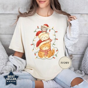 May include: An ivory short-sleeve t-shirt featuring a graphic of three stacked cats wearing red Santa hats and red plaid scarves, surrounded by a string of colorful Christmas lights. A circular label on the shirt displays the text IVORY.